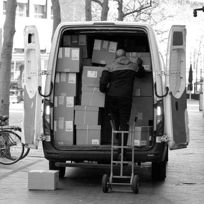 A person from Man with Van Hornsey is seen loading numerous cardboard boxes into the open rear of a large white van on a paved urban street. The individual, dressed in a dark jacket with a lighter upper section, is standing inside the vehicle, reaching up to arrange or place boxes among others stacked on the floor and shelves within the van. Several boxes have visible labels and barcodes, indicating packed goods for a home relocation or moving service. A small hand truck or dolly is positioned nearby, ready for transport of other items, and a single cardboard box rests on the ground just outside the van's open door. To the left, a bicycle is parked against a lamppost, and in the background, tree-lined streets and modern buildings are visible, suggesting an outdoor loading area on a clear day. This scene depicts the logistics involved in furniture transportation and packing during a professional moving process connected to house removals, handled by Man with Van Hornsey.