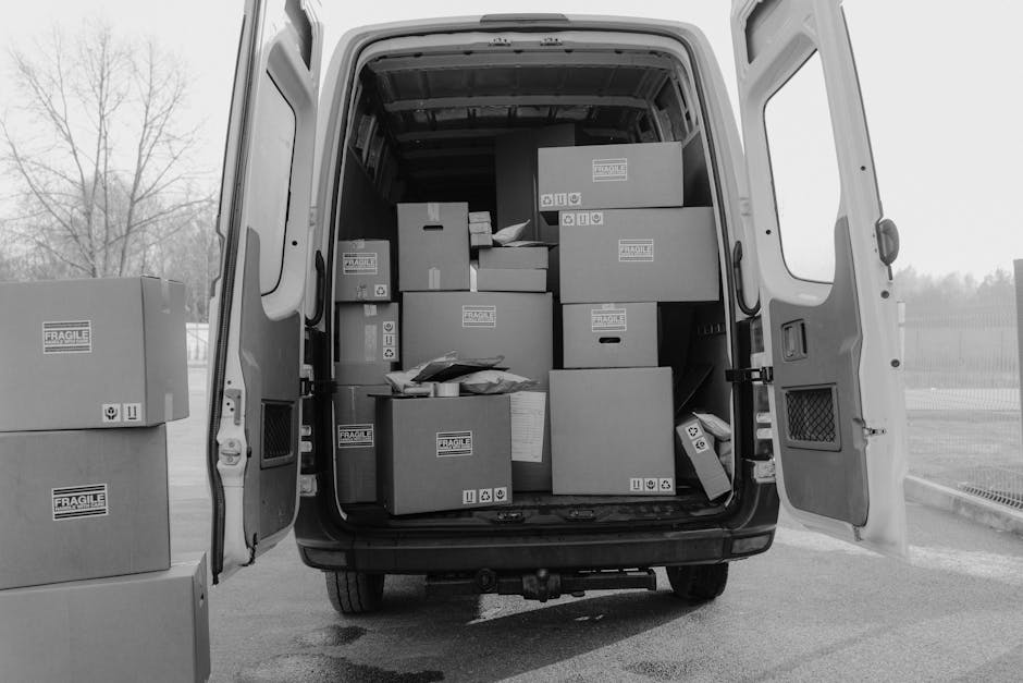 Inside the open rear of a delivery van, numerous cardboard boxes of varying sizes are stacked on the van's floor and shelves, some labeled with 'FRAGILE' stickers. Several boxes are positioned on the pavement outside the van, awaiting loading or unloading. The boxes are secured with packing materials such as paper and plastic wrap to ensure safe transport during home relocation or furniture transport. The van is parked on a paved surface adjacent to an outdoor area with a metal fence and leafless trees visible in the background, indicating a likely cold season. The image captures the loading process managed heightening the efficiency of packing and moving operations, with the focus on the careful handling of boxes within the vehicle, consistent with house removal services provided by [COMPANY_NAME]. Natural daylight illuminates the scene, emphasizing the organized packing as part of the logistics for a professional removal or relocation service in N8, Hornsey.