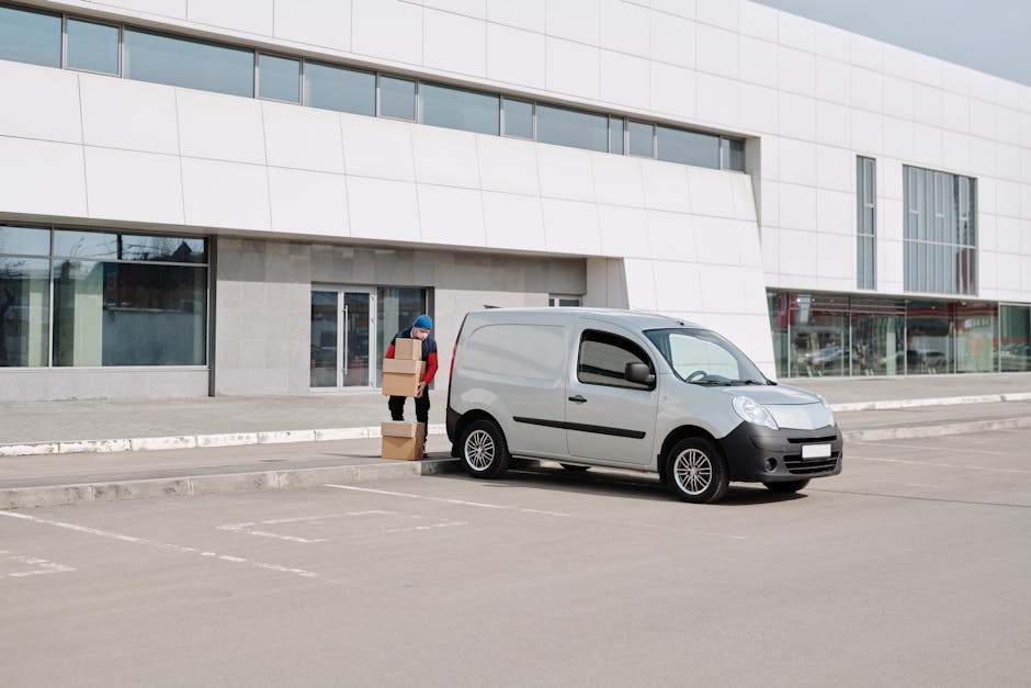 A person from Man with Van Hornsey is seen loading numerous cardboard boxes into the open rear of a large white van on a paved urban street. The individual, dressed in a dark jacket with a lighter upper section, is standing inside the vehicle, reaching up to arrange or place boxes among others stacked on the floor and shelves within the van. Several boxes have visible labels and barcodes, indicating packed goods for a home relocation or moving service. A small hand truck or dolly is positioned nearby, ready for transport of other items, and a single cardboard box rests on the ground just outside the van's open door. To the left, a bicycle is parked against a lamppost, and in the background, tree-lined streets and modern buildings are visible, suggesting an outdoor loading area on a clear day. This scene depicts the logistics involved in furniture transportation and packing during a professional moving process connected to house removals, handled by Man with Van Hornsey.