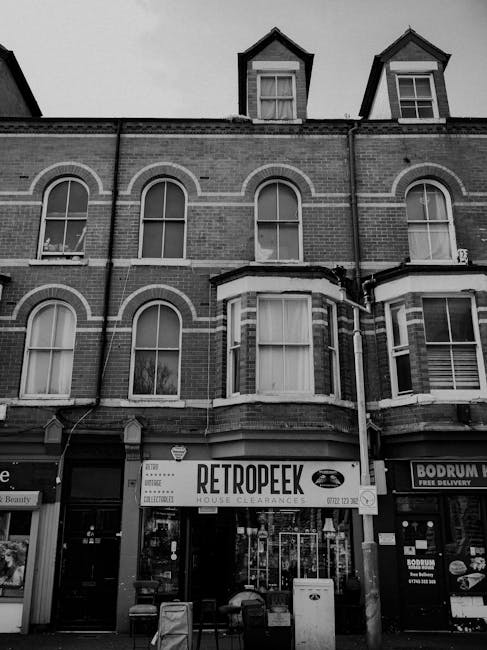 A street scene showing a collection of furniture and household items organized outside a row of shops, including a sofa, armchair, and several cardboard boxes, all prepared for moving. The items are p