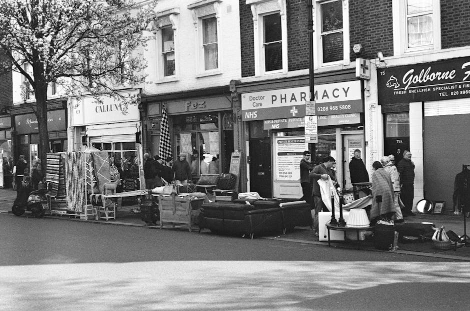A busy street scene in Hornsey featuring a temporary outdoor market setup with various furniture items and household goods displayed for sale, including sofas, chairs, and tables arranged along the pavement in front of a row of shops. Several shoppers and vendors are gathered around, some browsing and others engaged in conversation. The shops include a pharmacy with a sign reading 'Doctor Care NHS' and other retail outlets. A van is parked nearby, partially visible on the right side of the image, suggesting transportation or loading activities related to house removals or home relocation services. The environment is well-lit, with trees along the sidewalk providing partial shade. The scene captures typical urban activity associated with furniture transport and packing in preparation for moving, which aligns with the services offered by [COMPANY_NAME] as part of their removals and logistics solutions.