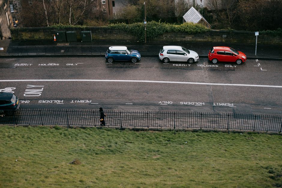 An aerial view of a street with three parked cars in designated permit holder parking bays, including a silver hatchback, a blue car with a white roof box, and a red vehicle, positioned behind a black metal fence separating the parking area from a grassy verge. The street surface displays painted markings indicating 'Permit Holders Only,' with some interruption by a white arrow directing traffic flow. A pedestrian, dressed in dark clothing, is walking along the pavement next to the fence. In the background, there is a row of residential buildings and trees, with a small orange traffic cone placed near the curb. This scene illustrates typical urban parking provisions and showcases the parking environment for house removal services like those offered by Man with Van Hornsey, which may involve loading and unloading furniture and boxes from vehicles, utilizing suitable equipment to facilitate home relocation processes.