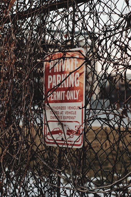 A metal wire fence with intertwined dry, brown vines predominantly covering it, obscuring parts of the view behind. Attached to the fence is a red and white parking permit sign that reads 'PRIVATE PARKING PERMIT ONLY,' with additional text indicating restrictions on unauthorized vehicles. The sign is positioned near the top center of the image and appears slightly weathered. The background features a blurred outdoor scene with a pale sky and some distant structures, suggesting an open area or roadside setting. This environment aligns with typical surroundings for house removals or relocation services, where parking permits may be necessary for loading or unloading parked vehicles, such as trucks or vans. The image emphasizes the importance of parking compliance during home relocations conducted by companies like Man with Van Hornsey, ensuring smooth and lawful furniture transport preparations.