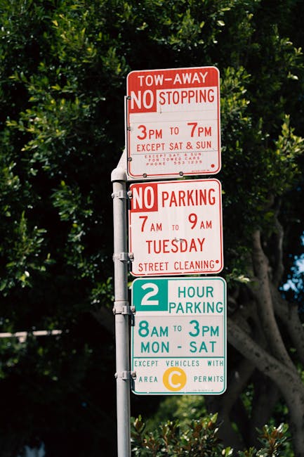 An aerial view of a street with three parked cars in designated permit holder parking bays, including a silver hatchback, a blue car with a white roof box, and a red vehicle, positioned behind a black metal fence separating the parking area from a grassy verge. The street surface displays painted markings indicating 'Permit Holders Only,' with some interruption by a white arrow directing traffic flow. A pedestrian, dressed in dark clothing, is walking along the pavement next to the fence. In the background, there is a row of residential buildings and trees, with a small orange traffic cone placed near the curb. This scene illustrates typical urban parking provisions and showcases the parking environment for house removal services like those offered by Man with Van Hornsey, which may involve loading and unloading furniture and boxes from vehicles, utilizing suitable equipment to facilitate home relocation processes.