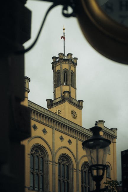 A view of Hornsey Town Hall's historic clock tower and ornate facade, seen through a window with a vintage-style street lamp and part of a clock mechanism at the top of the image. The building features arched windows, decorative stonework, and a flag flying from the tower's flagpole. Overcast sky provides diffuse lighting, and the scene captures the exterior architecture of the town hall, which is a common landmark in house and office relocation contexts. Man with Van Hornsey occasionally manages moving logistics around such buildings, supporting access, parking, and timing for home removal projects.