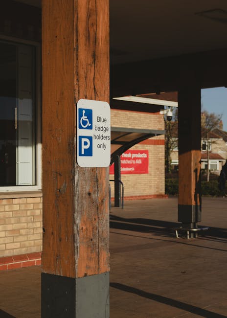 A white parking sign attached to a wooden post outside a brick building indicates that the area is reserved for blue badge holders only, featuring a blue wheelchair symbol and a large blue 'P' for parking. The wooden post is part of an outdoor covered area with a pavement underneath, leading to a spacious parking lot or driveway. In the background, a building with a red sign, windows, and a residential setting with houses and trees are visible under clear daylight. This scene suggests the location is near a public or community facility, possibly related to house removals or moving services, where careful access and parking arrangements are important for loading or unloading furniture and packing materials during home relocation activities, supported by companies like Man with Van Hornsey.
