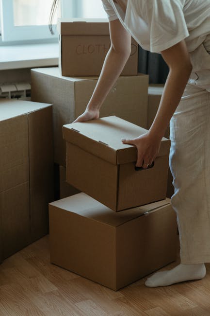 A person wearing a white shirt and light-colored trousers is seen moving and stacking cardboard boxes inside a home, preparing for a house relocation. One box is being placed on top of other boxes on