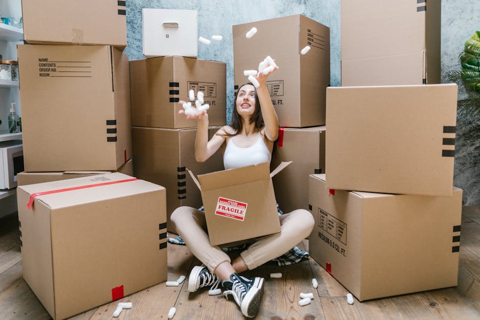 A person wearing a white shirt and light-colored trousers is seen moving and stacking cardboard boxes inside a home, preparing for a house relocation. One box is being placed on top of other boxes on