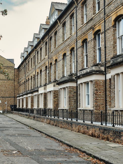 A row of traditional terraced houses on Hornsey High Street, with brick facades and white-framed windows. The pavement in front features a low, black metal railing separating the sidewalk from the building entrances. The ground-floor windows have white shutters or blinds, and the upper floors display small dormer windows with slate roofing. On the narrow street, there are various cardboard boxes, wrapped furniture, and plastic packing materials placed next to the house entrances, indicating an active home relocation process. Scattered leaves are visible on the pavement, and an orange van is partially visible at the far end, suggesting transportation of household items by a professional removals service, such as Man with Van Hornsey. The scene is lit with natural daylight, capturing the preparation and logistical aspects of furniture transport and packing involved in house removals onto the street front during a typical moving day in Hornsey.