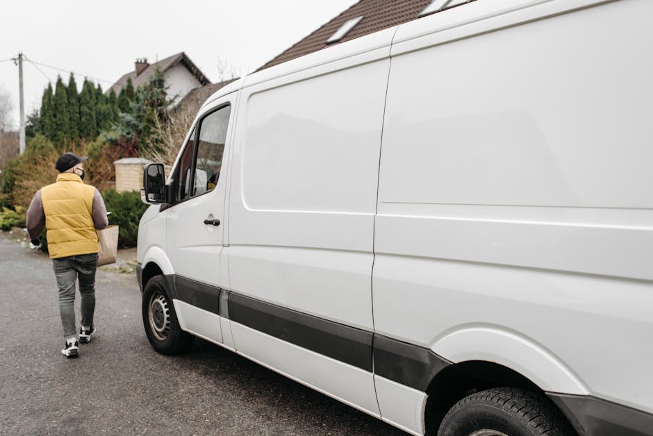 A man wearing a yellow insulated vest, grey trousers, and a black cap with glasses is walking on a residential street, carrying a cardboard box towards a white cargo van parked next to a driveway. The van, which is part of a home relocation or furniture transport service by Man with Van Hornsey, is positioned with its side door open on a paved road, close to the curb. Behind the van, there are suburban houses with sloped roofs, some trees, bushes, and a utility pole visible in the background. The scene is set during daylight with overcast weather. Inside the open van, partially visible, are packing materials such as bubble wrap and cardboard boxes, indicating a packing and moving process. The van appears to be in the midst of a loading operation, with the individual preparing to load or unload items as part of a professional removal or relocation service in north London.