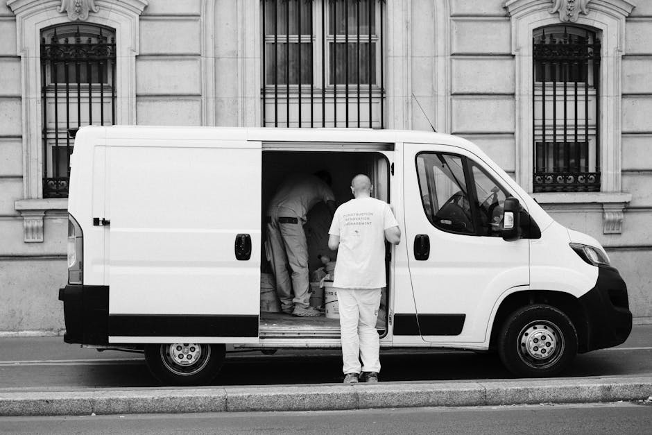 A white moving van parked on a street in front of a two-storey residential building with a brick and white exterior facade. The van displays the text 'MOVING COMPANY LOCAL & LONG DISTANCE' on its side. Inside the driver's cabin, a man wearing sunglasses and a cap is visible, engaging in a conversation with a man standing outside the van. The man outside, dressed in a dark t-shirt and trousers, stands with arms crossed, smiling, and facing the driver. The building features large arched windows with white frames and a brick detail surrounding the upper windows, indicating a typical residential house. The scene is set during daytime with clear weather, and the overall context suggests preparations for home relocation, with the men involved in loading or arranging furniture and boxes for transport by [COMPANY_NAME], specialists in house removals and furniture transport.