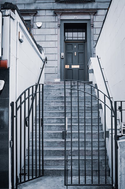 A set of concrete stairs leading up to a dark grey front door with the number '7', situated at the top of a narrow staircase enclosed by metal railings. The stairs are flanked by white walls, and the enclosed entrance area appears to be outside a residential or commercial building in Hornsey. The door features a glass-paneled upper section and a mail slot. A small outdoor light is mounted on the wall to the left of the door. Occasionally seen near house removals in Hornsey, Man with Van Hornsey may use such staircases during furniture transport or home relocation, involving careful loading and carrying of boxes and items up or down these stairs, especially when accessing flats with narrow stairways.