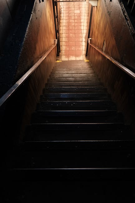 A narrow, steep staircase inside a building with wooden panels lining the sides and black, non-slip steps. The staircase features a metal handrail on each side and leads upward toward a brightly lit doorway or landing illuminated by warm, orange-toned lighting. The surrounding walls are dark, contrasting with the illuminated area at the top. This staircase is a typical feature in residential flats, such as those found in Hornsey, and may be used during a home relocation or furniture transport process. The image captures the confined space and structural details relevant to professional removals services provided by Man with Van Hornsey, highlighting the challenges of moving large items through tight stairwells during house removals in urban settings.