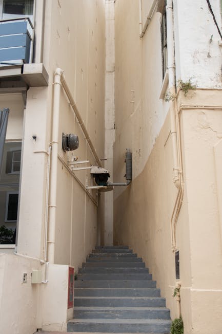 A set of concrete stairs leading up to a dark grey front door with the number '7', situated at the top of a narrow staircase enclosed by metal railings. The stairs are flanked by white walls, and the enclosed entrance area appears to be outside a residential or commercial building in Hornsey. The door features a glass-paneled upper section and a mail slot. A small outdoor light is mounted on the wall to the left of the door. Occasionally seen near house removals in Hornsey, Man with Van Hornsey may use such staircases during furniture transport or home relocation, involving careful loading and carrying of boxes and items up or down these stairs, especially when accessing flats with narrow stairways.