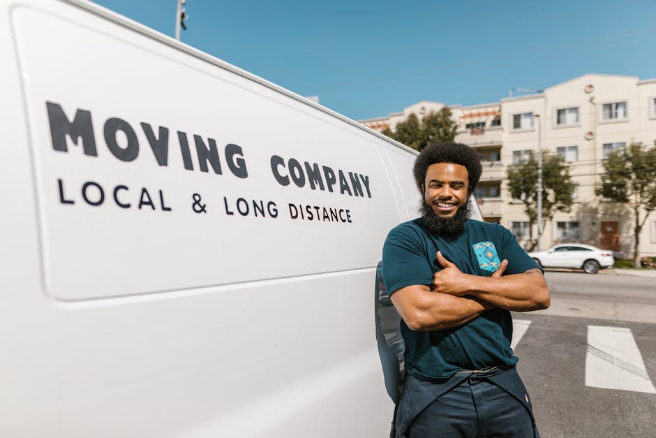 A smiling man with a full beard and curly hair stands confidently with arms crossed next to a white moving van parked on a street in Hornsey. The van displays the company name 'Moving Company' with the tagline 'Local & Long Distance' written underneath in bold black letters. The man is wearing a dark blue T-shirt with a small logo on the chest. In the background, residential buildings with multiple stories, balconies, and numerous windows are visible, along with parked cars, trees, and a clear blue sky. The scene appears to be during daytime with natural lighting highlighting the vehicle and the man. This image illustrates home relocation or furniture transport services, emphasizing professional moving logistics and packing processes provided by [COMPANY_NAME], as they prepare for an efficient move within or beyond Hornsey.