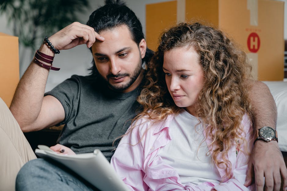 A man and a woman sitting closely together on a sofa inside a home, surrounded by cardboard boxes indicative of a house move. The man, with dark hair and a beard, is wearing a grey t-shirt and has a thoughtful expression while resting his head on his hand, which is supported by his elbow. The woman, with curly blonde hair, is wearing a light pink shirt and appears concerned or focused as she looks at a tablet in her hands. In the background, there are additional stacked moving boxes, some with visible labels, and a doorway leading to another room. The environment suggests an ongoing packing and moving process, with visible packing materials such as cardboard boxes and protective wrapping on items, reflecting typical furniture transport and logistical aspects of home relocation handled by services like Man with Van Hornsey.